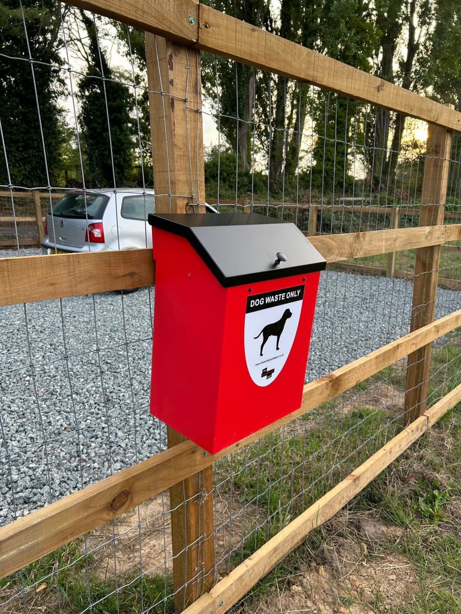 A red dog waste bin mounted on the fence beside the parking area